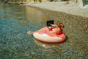 Woman laptop sea. Freelancer woman in sunglases floating on an inflatable big pink donut with a laptop in the sea. People summer vacation rest lifestyle concept