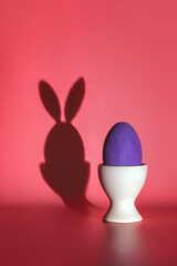 purple Easter egg on a stand, shadow of an egg with bunny ears on the wall, creative Easter card, minimalistic style, pink background