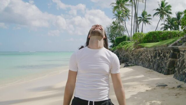 Young handsome bearded tourist in white T-shirt laughing, touching gently his fair hair standing on sunny shore with palm trees of Hawaii island, Oahu. High quality 4k footage