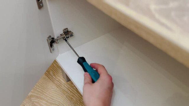 A worker is screwing a hinge into a wooden cabinet door with a screwdriver. Adjustment of fittings, door hinges. Home renovation. Furniture assembly.