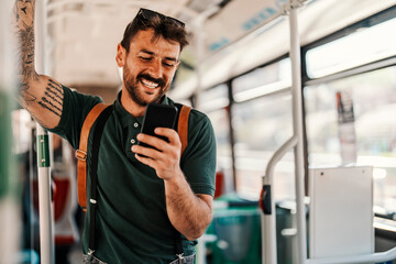 A trendy passenger is scrolling on the phone and smiling at it while riding a bus.