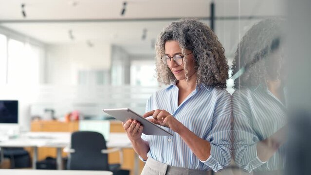 Mature busy happy businesswoman bank manager, older female corporate executive holding digital tablet standing at work. Middle aged professional business woman using tab computer in office.