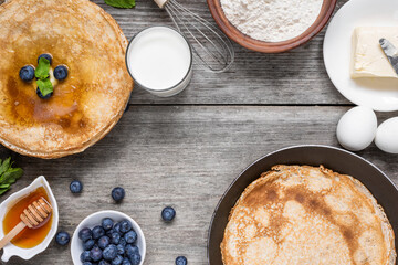 Stack of thin pancakes with blueberries and honey and frying pan with delicious crepes and ingredients for cooking on rustic wooden table. Top view. Delicious breakfast