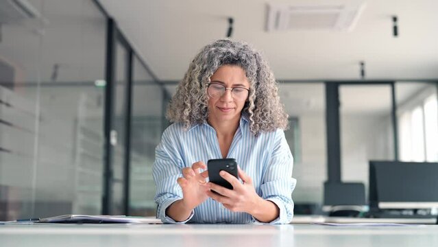 Busy mature professional businesswoman executive using smartphone cell mobile apps on cellphone working sitting at desk. Middle aged business woman holding phone using cellphone in office.