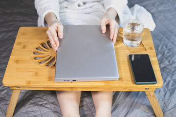 Woman is Working with laptop, smartphone and glass of water on the bed. Modern gray thin laptop on the wooden Cooling pad for laptop. Notebook Cooler for Computer, USB Fan.