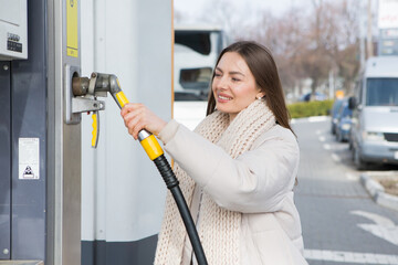 Young woman holding a fuel nozzle in her hand while refueling car at gas station. A stop for...