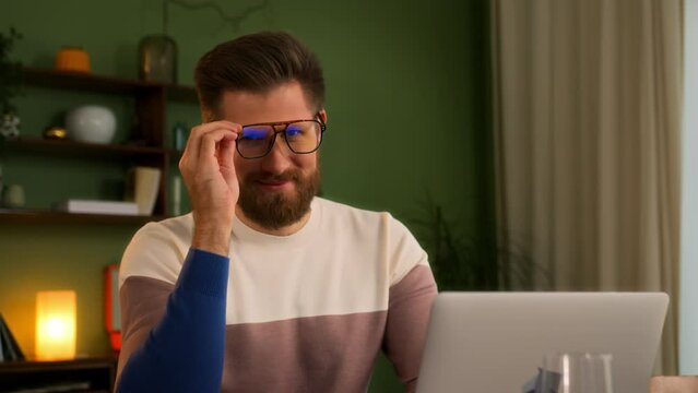 portrait young contented man with beard and eyeglasses sits at table indoors and works on laptop typing on keyboard. pleased ambitious student on online education. remote specialist advanced training.