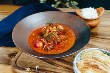 A bowl of red curry with meat and vegetables sits on a wooden table