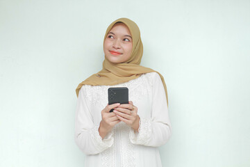 Young Asian Muslim woman holding mobile phone with happy expression with thinking something. Isolated in gray background.