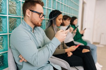 Side view of focused young diverse ethnicity candidates using mobile phones while waiting job interview in modern office lobby. Bored male and female for vacancy sitting on chairs in queue corridor.