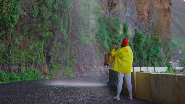 Woman traveler hiker in a yellow raincoat near waterfall in the highlands, enjoys nature and life. Traveling in the mountains, adventure in trip. Lifestyle concept. Traveller in yellow puffer jacket