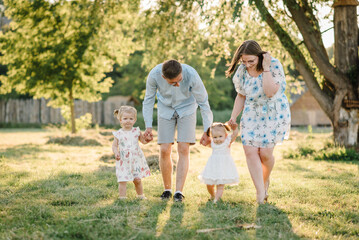 Obraz premium Mom, dad, and kids walking in park at sunset. Mother, father hold hands happy children daughter. Running in green grass in nature on summer day. Concept of family spending time together on a picnic.