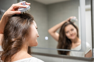 Fototapeta premium Young woman doing self hair scalp massage with scalp massager or hair brush for hair growth stimulating at home. Reflected view of the mirror