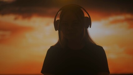Silhouette of a woman listening to music on large wireless headphones against a sunset background