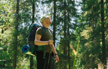 Adult man with backpack and hiking stick walking in green forest in daylight