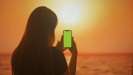 Silhouette of a woman viewing information on a smartphone with green screen at sunset