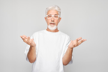 Senior man with gray hair in glasses and white shirt making uncertain gesture with hands raised