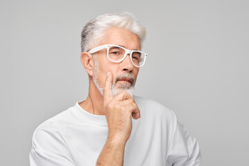 Pensive senior man with white hair and glasses, hand on chin, isolated on grey background.