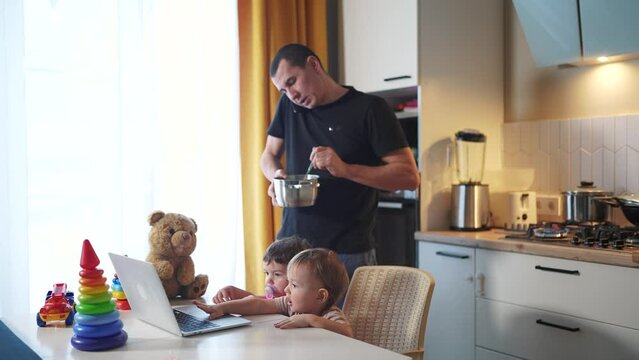 father cooking in the kitchen remote work. baby twins playing with laptop in the kitchen. father working at home in the kitchen lifestyle stirring food in the pan. remote work business concept