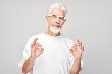 Senior man with glasses making OK gesture on a gray background.
