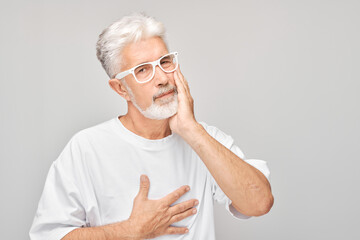 Elderly man with glasses touching cheek in discomfort, dental pain, against a gray background.