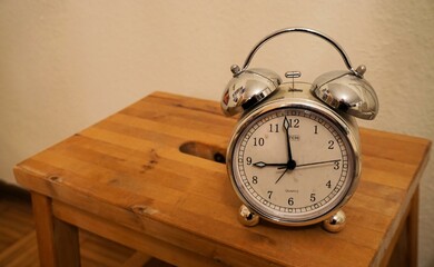 Silberner Wecker mit Glöckchen steht auf braunem Nachttisch aus Holz vor weißer Wand in...