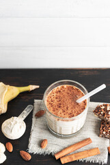 Glass with protein drink, healthy milkshake smoothie on wooden board with bananas, protein powder in measuring spoon, protein bar, almond nuts and cinnamon sticks