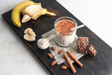 Glass with protein drink, healthy milkshake smoothie on wooden board with bananas, protein powder in measuring spoon, protein bar, almond nuts and cinnamon sticks