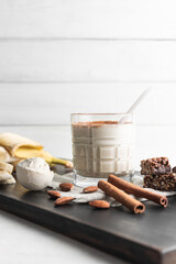 Glass with protein drink, healthy milkshake smoothie on wooden board with bananas, protein powder in measuring spoon, protein bar, almond nuts and cinnamon sticks
