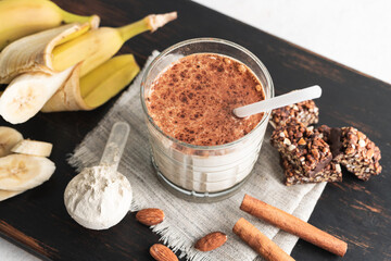 Glass with protein drink, healthy milkshake smoothie on wooden board with bananas, protein powder in measuring spoon, protein bar, almond nuts and cinnamon sticks
