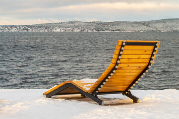 A wooden beach chair is sitting on the snow next to a body of water. The chair is empty and the...