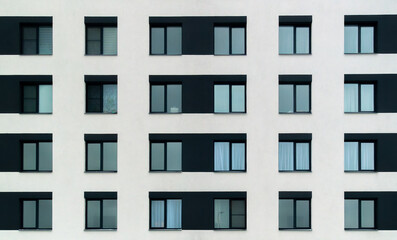 Fototapeta premium The building is tall and has many windows. The windows are mostly black and white, with some blue accents. The building appears to be a modern apartment building with a minimalist design