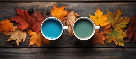 Two mugs filled with tea are placed on a wooden table, surrounded by a variety of colorful autumn leaves.