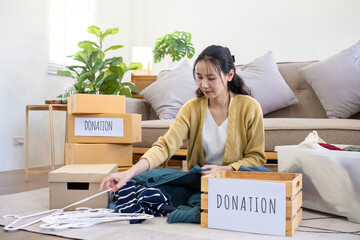 Cute Asian woman sitting next to sofa in living room sorting unwanted clothes for donation.