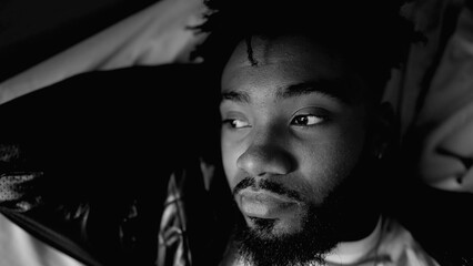 Contemplative young African American man lying in bed gazing while in deep mental reflection,...