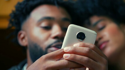 African American young couple close-up faces staring at cellphone device in foreground. People watching entertainment media on phone screen, consuming entertainment media