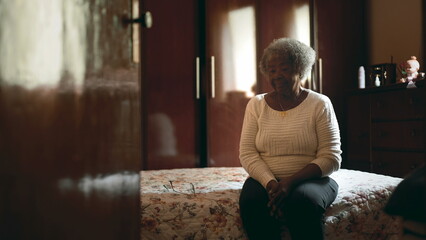 Candid Senior South American black lady with gray hair sitting by bedside with contemplative gaze pondering old age and life's challenges, humble home background