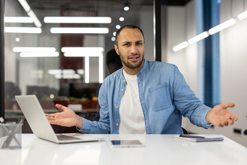 Disappointed and upset young hispanic man in blue shirt sitting at desk with laptop in office,...