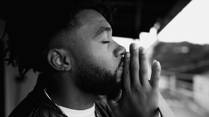 Hopeful young man PRAYING to GOD with eyes closed and hands clenched together UPWARDS. Faithful and...