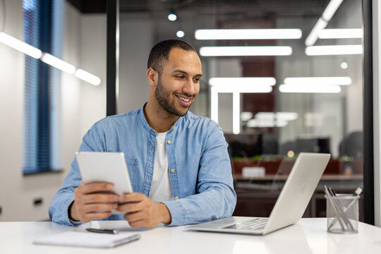 A young Hispanic man is talking on a video call on a laptop, sitting at a desk in the office, looking at the screen with a smile and holding a tablet in his hands - Powered by Adobe
