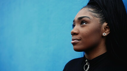 One contemplative black latina girl gazing upwards with HOPE and FAITH leaning on blue wall in urban street environment. African American young woman portrait looking up with thoughtful gaze