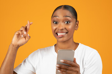 Hopeful black woman poses crossing fingers while using smartphone, studio