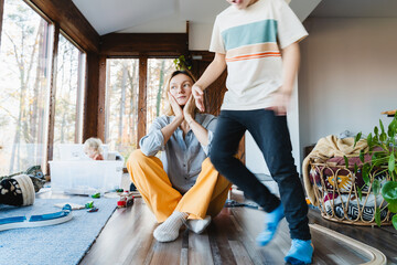 Stressed out mother sitting in middle of toys while children naughty running around her at room.