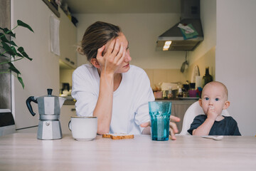 Exhausted young woman with baby is sitting with coffee in kitchen. Modern tired mom and little...