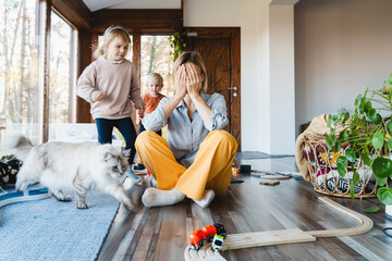 Stressed out mother sitting in middle of toys while children naughty running around her at room.