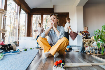 Stressed out mother sitting in middle of toys while children naughty running around her at room.