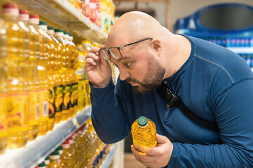Shocked overweight man raising his glasses to his forehead and looking on price of vegetable oil in...