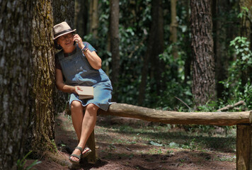 A woman is sitting and leaning against a tree trunk while talking on the phone; a book is in her lap