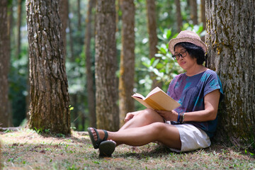 A woman is sitting under a tree while reading a book; serious expression - obrazy, fototapety, plakaty
