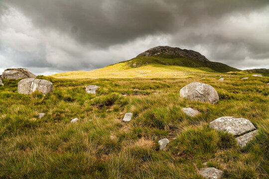 Visit Ireland: sunlight and cloud shadows over a stony outcrop rising from yellow grass on a mountainside in the Connemara National Park